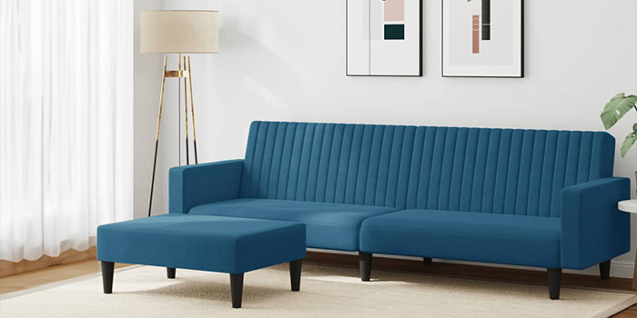 Moderna plava baršunasta kutna garnitura s okomitim prošivima i odgovarajućim tabureom. Sofa ima crne drvene noge i smještena je u svijetlom dnevnom boravku s uokvirenim slikama na zidu i podnom lampom u kutu.